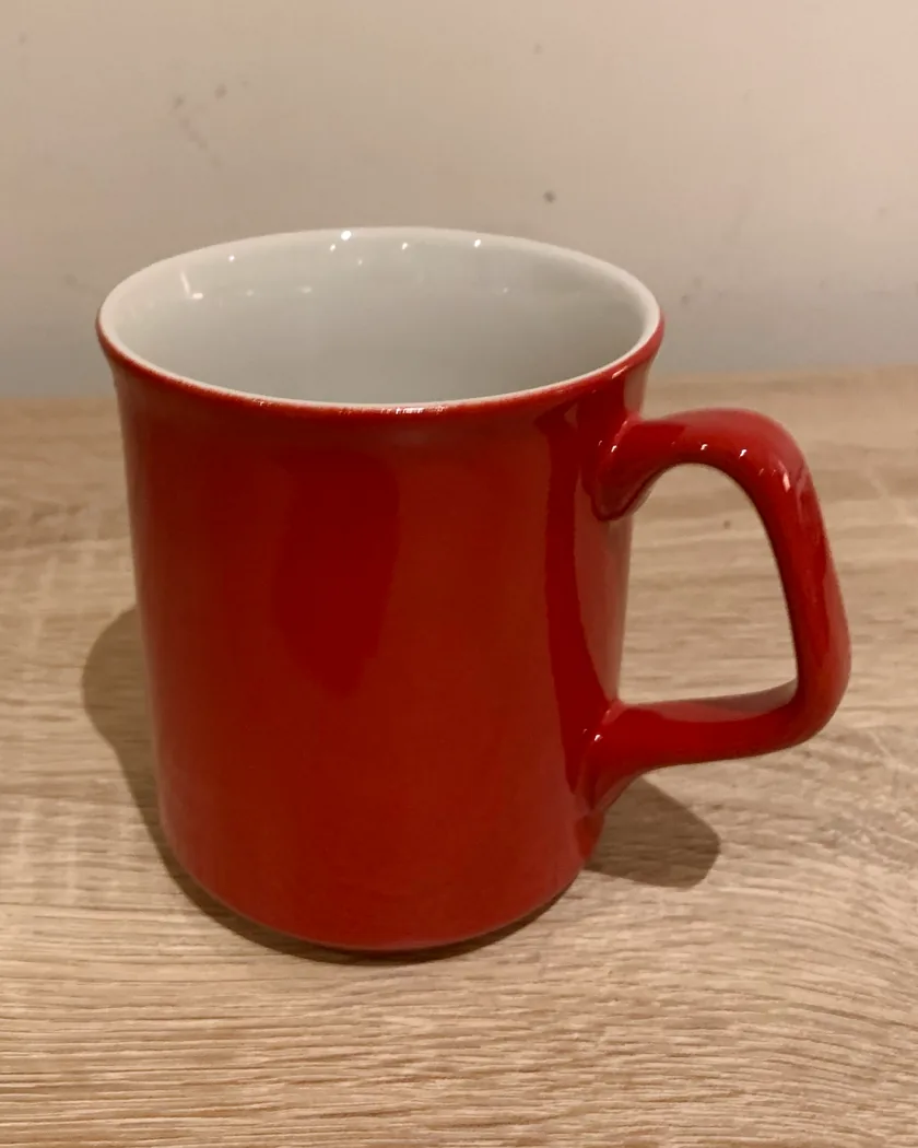 Lot 001, the actual mug: a glossy red mug with a white inside and an angular handle on the right, standing on a pale oak desk.